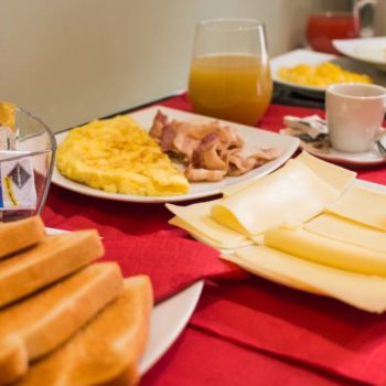 Dettaglio colazione, a sx fette di pane tostato, vaso contenente marmellate in monoporzione, al centro piatto con fette di formaggio, piatto con omelette affiancata da bacon, a dx tazzina caffè espresso e una caraffa di succo