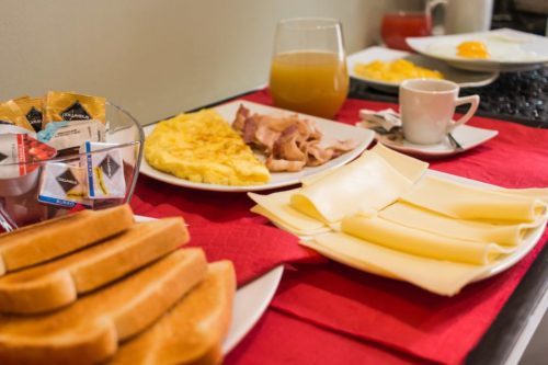 Detail breakfast, left slices of toast, jar containing jam in single portion, in the center plate with slices of cheese, dish with omelet flanked by bacon, right cup espresso coffee and a carafe of juice